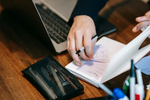 Man's hands writing notes in a notebook beside a laptop on a wooden desk. Hitta rätt tjänstekategori för ditt behov